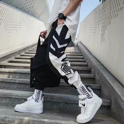 Black and white jogger pants with bold geometric designs, paired with white sneakers and a casual tee, showcasing trendy streetwear fashion on modern concrete stairs.